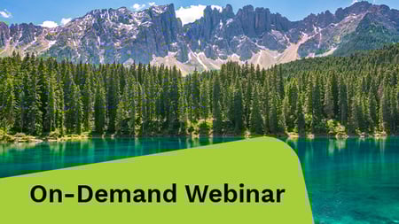 Bright turquoise alpine lake reflects a dense evergreen forest backed by rugged, partially snow-capped mountains under a blue sky.