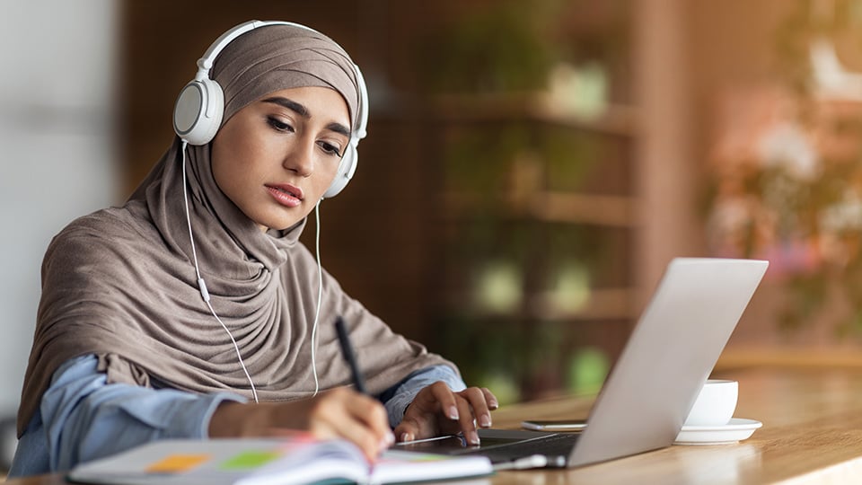 Girl in headscarf having online lesson on laptop at cafe, wearing headset, taking notes, online education concept, copy space