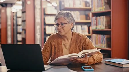 Senior woman, computer and planning in a library with book and laptop for lecture class.