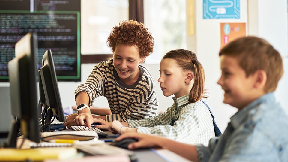 Group of kids sitting at desks using desktop computers in classroom, one curly haired boy helping classmates with programming tasks, monitors displaying code on screens