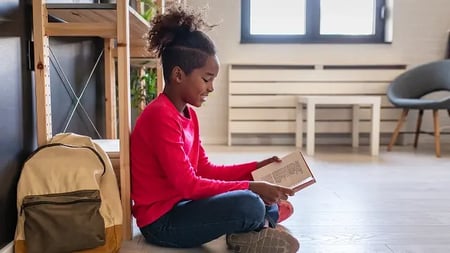 Student sitting on the floor near a backpack, reading a book in a bright indoor space with natural light from a window