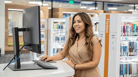 A smiling woman checks the library computer to find a book title.