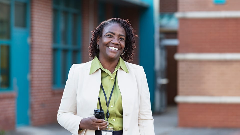 A mature African-American woman standing outside a school building, smiling and looking away from the camera. She is a teacher or school principal.