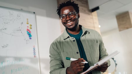 District tech coordinator happily smiles in a modern meeting room, holding a notebook and marker near a whiteboard