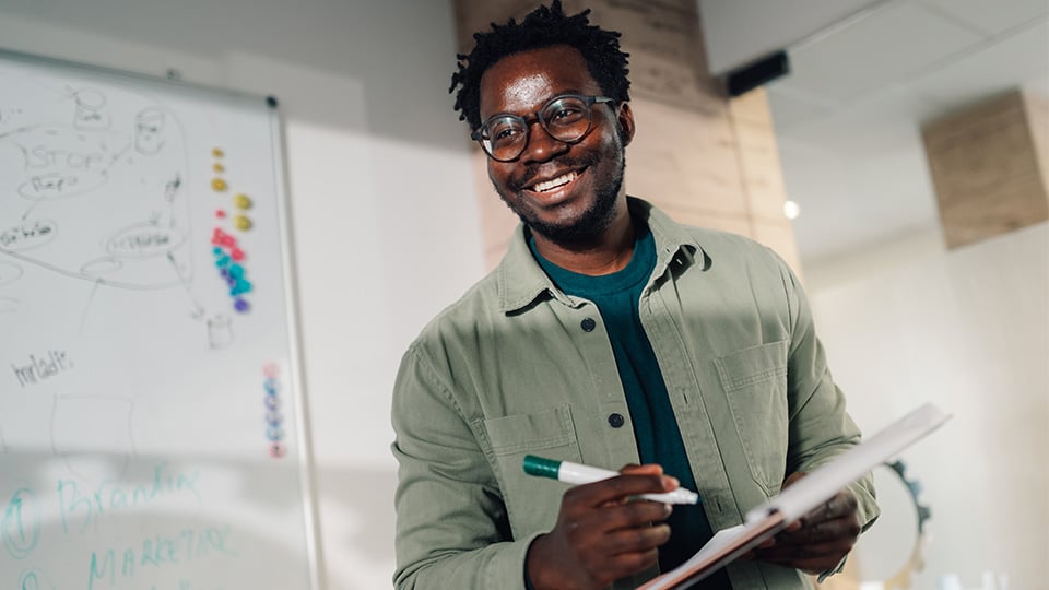 District tech coordinator happily smiles in a modern meeting room, holding a notebook and marker near a whiteboard